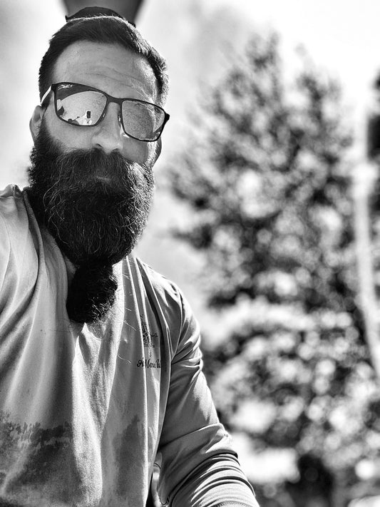 Bearded man wearing glasses and a long-sleeve shirt, standing outdoors near a pine tree, looking confident and bold.