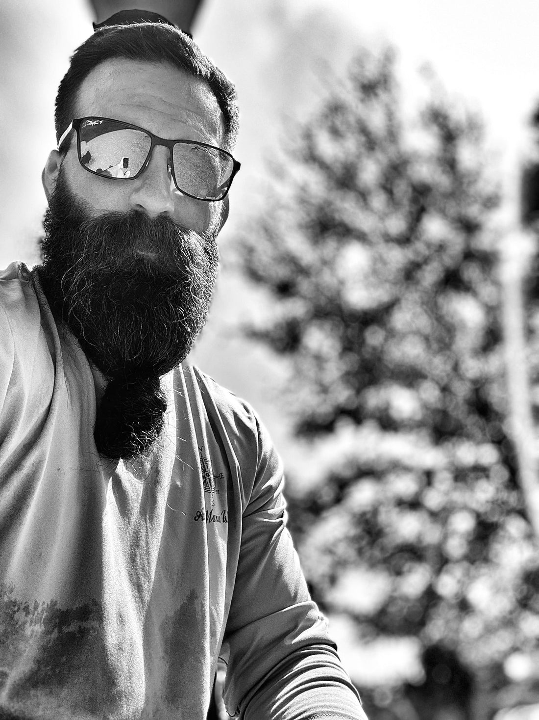 Bearded man wearing glasses and a long-sleeve shirt, standing outdoors near a pine tree, looking confident and bold.